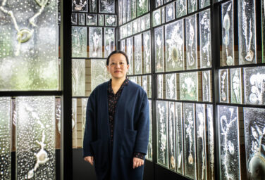 Rui Sasaki, an artist based out of Japan, standing in front of a structure that has glass panes as walls, each pane being an impression of a plant.