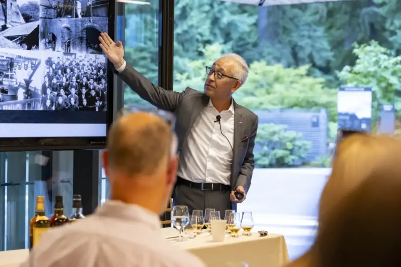 Suntory Executive Officer and Chief Blender, Shinji Fukuyo stands in front of several glasses of whisky and gestures to a presentation on his company on a nearby monitor.