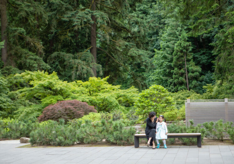 Adult woman and girl sitting on bench among the green foliage within the Japanese Garden.
